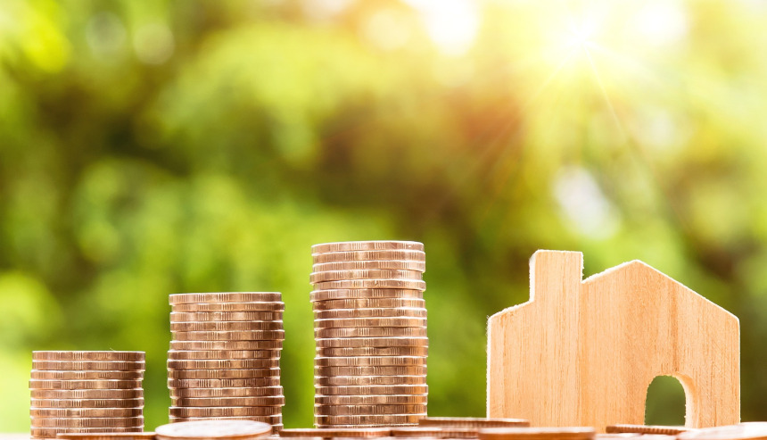 Stacks of coins and a wooden house on a table with a green background and sunlight