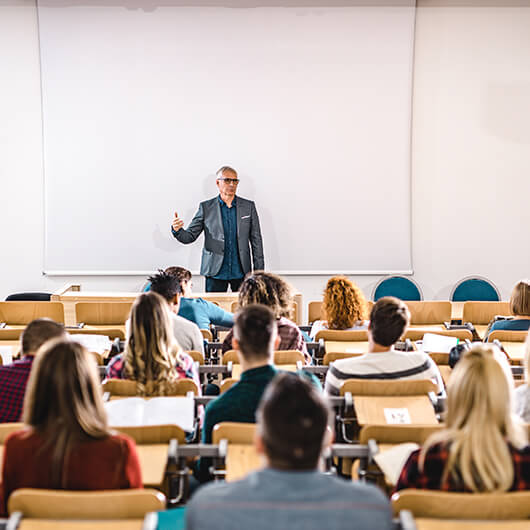 A male teacher with glasses is lecturing to students in an auditorium