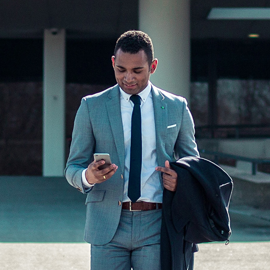 a man in a suit is walking in front of the big office building writing somthing on his phone and holding his jacket with another hand